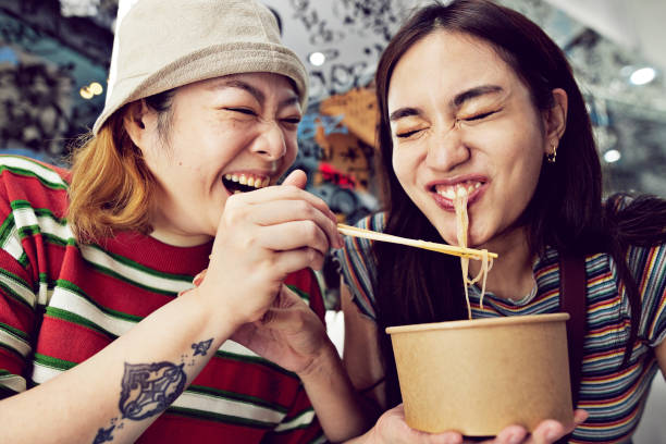 happy young woman feeding noodles with chopsticks to female friend at street